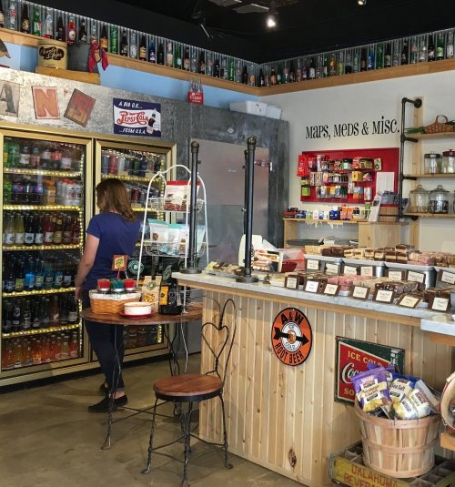 A cozy store interior with shelves of snacks, drinks, and vintage decor. A person browses the soda selection.