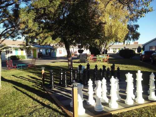 A large outdoor chess set with black and white pieces, surrounded by trees and colorful chairs in a grassy area.