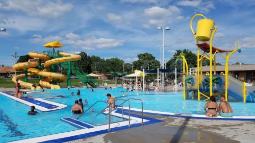 A vibrant outdoor pool scene with water slides, a splash pad, and people enjoying the water on a sunny day.