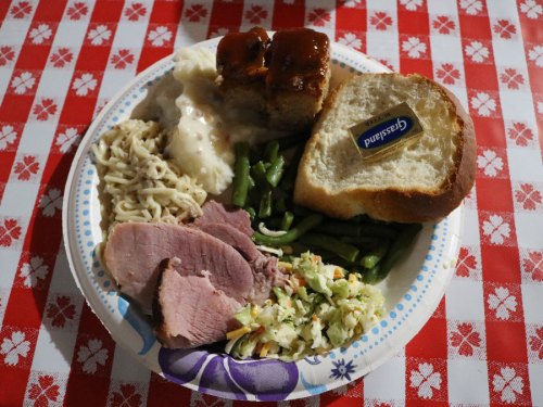 A paper plate with sliced ham, green beans, coleslaw, noodles, mashed potatoes, meatloaf, and a slice of bread.