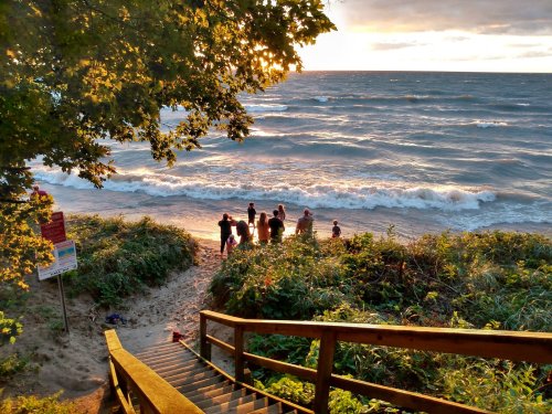 A group of people stands by the shore at sunset, with waves lapping at the beach and trees framing the scene.