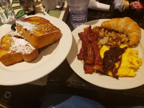 A plate with French toast dusted with powdered sugar and a plate with bacon, scrambled eggs, hash browns, and a croissant.