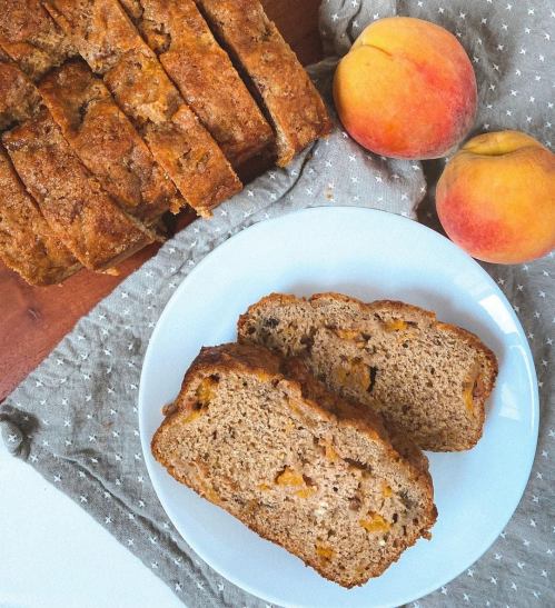 Sliced peach bread on a white plate, with whole peaches and a loaf in the background on a textured cloth.