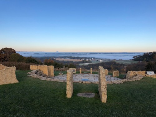 Ancient stone circle on a grassy hill, overlooking a misty valley and distant hills under a clear blue sky.