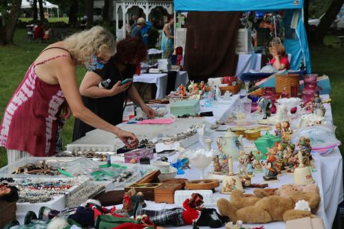 Two women browse a table filled with various vintage items and collectibles at an outdoor market.