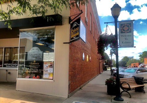 A street view of Kalona's storefront with a sign, hanging flowers, and a welcome banner in a charming town setting.