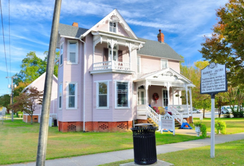 A large pink Victorian house with a porch, surrounded by grass and trees, featuring a historical marker nearby.