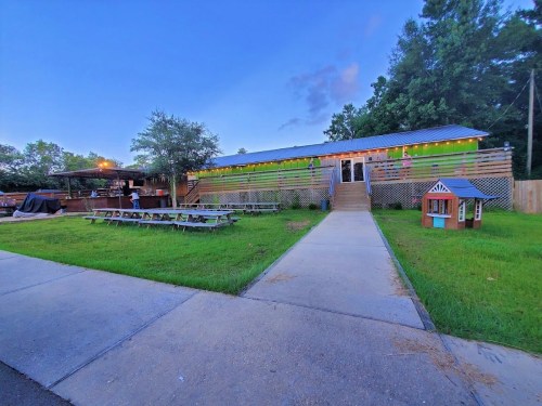 A rustic restaurant with outdoor seating, surrounded by greenery, under a twilight sky.