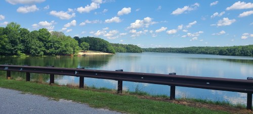 A serene lake surrounded by lush greenery and blue skies with fluffy clouds, viewed from a gravel path.