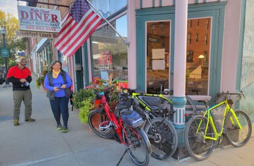 A woman in a blue jacket stands outside a diner with bicycles parked nearby and an American flag hanging above.