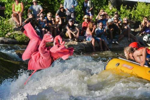 A person in a pink outfit flips into a river while spectators watch from the shore, enjoying a lively outdoor event.