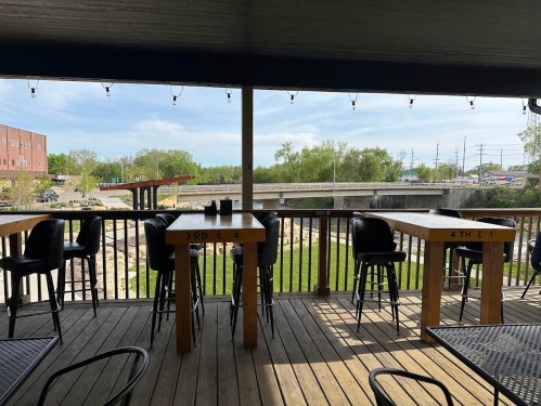 View from a deck with wooden tables and chairs overlooking a green area and a river, under a clear blue sky.