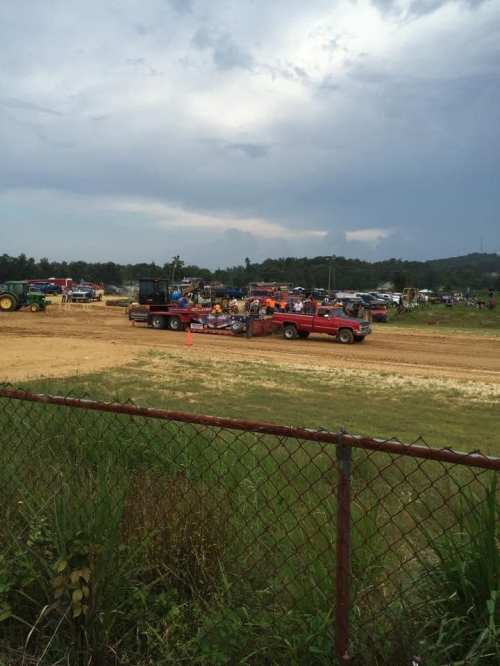 A rural scene with trucks and tractors at an outdoor event, surrounded by spectators and a cloudy sky.