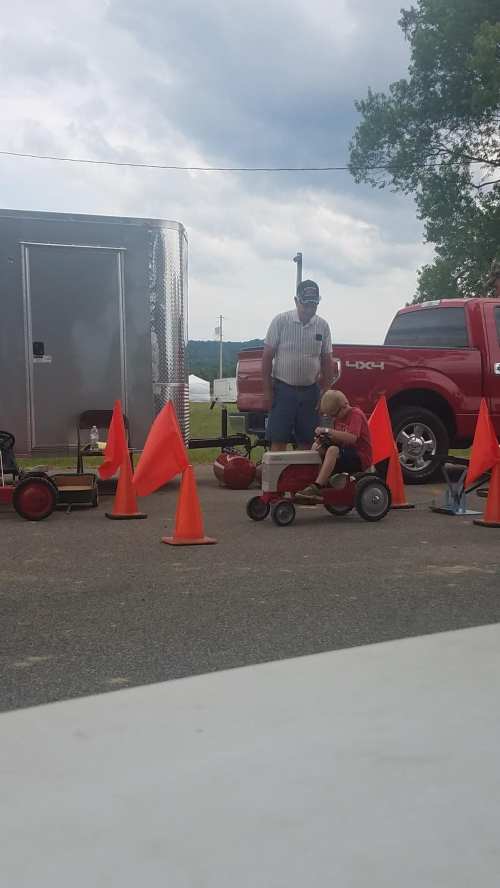 A man watches a child driving a small tractor among orange cones, with a red truck and trailer in the background.