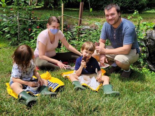 A family enjoys a sunny day in the garden, with two children sitting on the grass and parents nearby.