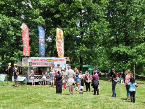 A busy food truck area in a park, with people enjoying funnel cakes and corn dogs under colorful banners.