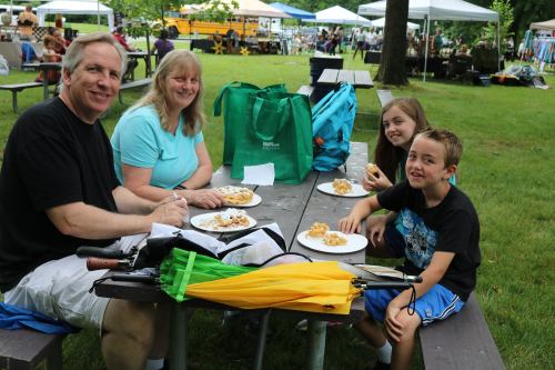 A family enjoys a meal at a picnic table in a park, surrounded by tents and greenery.