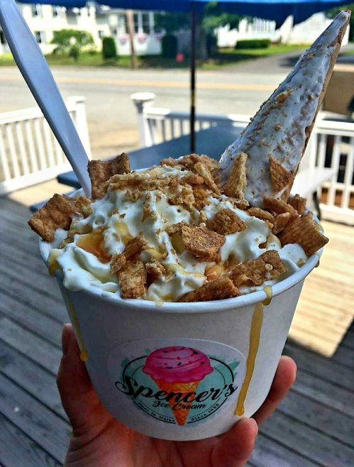 A hand holds a cup of ice cream topped with cereal, whipped cream, and a cone, with a sunny outdoor background.