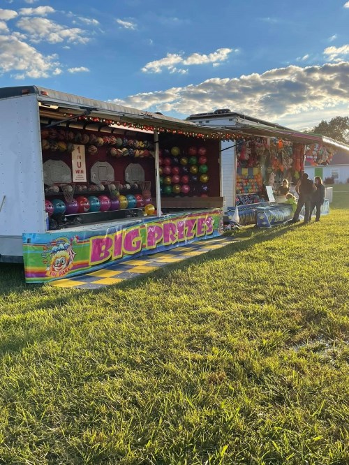 A colorful carnival game booth with prizes, set against a sunny sky and grassy field. People are playing games nearby.