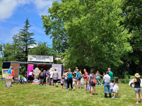 A crowd of people lines up at an ice cream shop in a grassy area, surrounded by trees and a blue sky.