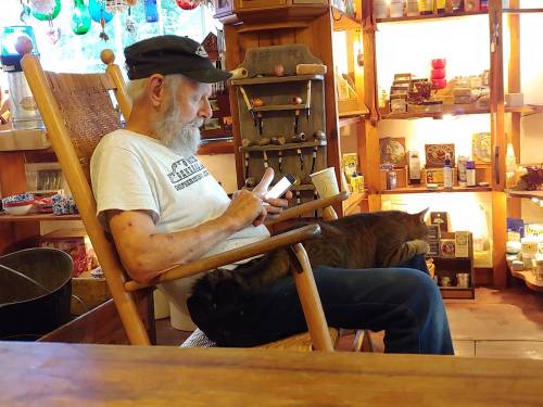 An elderly man in a rocking chair uses his phone, with a cat resting on his lap, surrounded by shelves of colorful items.