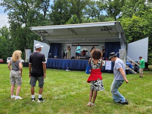 A small crowd watches a performance on an outdoor stage surrounded by trees on a sunny day.