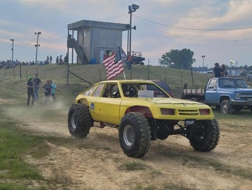 A yellow modified car with large tires drives on a dirt track, with spectators and a flag in the background.