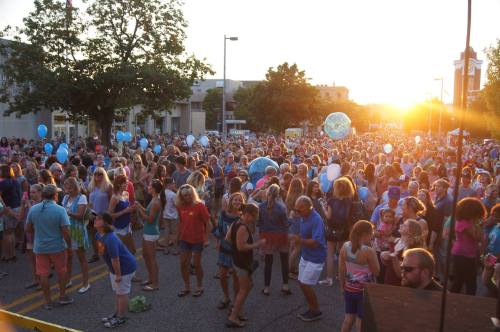 A large crowd gathers outdoors at sunset, enjoying a festive atmosphere with balloons and music.