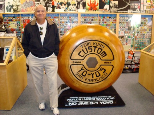 A man stands next to a giant wooden yo-yo display in a colorful shop filled with various yo-yos and memorabilia.