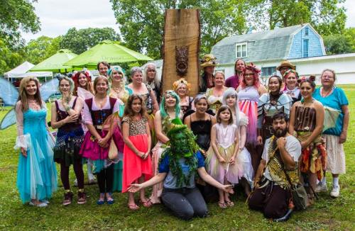 A diverse group of people in colorful costumes poses together outdoors, surrounded by greenery and tents.