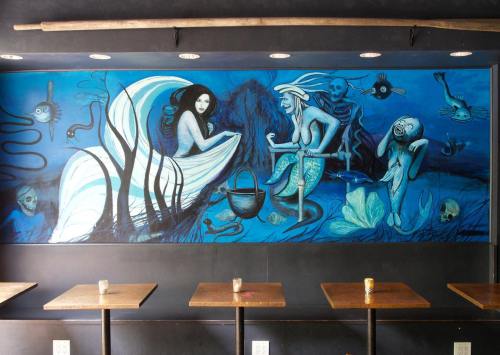 A vibrant mural featuring mermaids, sea creatures, and skulls in shades of blue, set against a dark wall in a café.