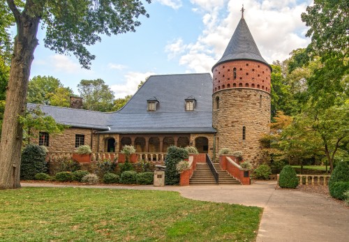 A stone building with a turret, surrounded by trees and greenery, under a partly cloudy sky.