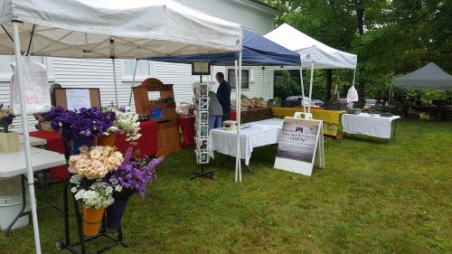 A vibrant outdoor market with tents displaying various goods, surrounded by greenery and colorful flower arrangements.