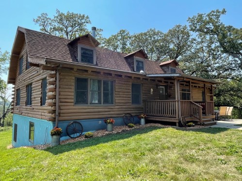 A charming log cabin with a porch, surrounded by greenery and trees under a clear blue sky.