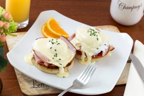 Two eggs Benedict on English muffins with ham, topped with hollandaise sauce and chives, served with orange slices.