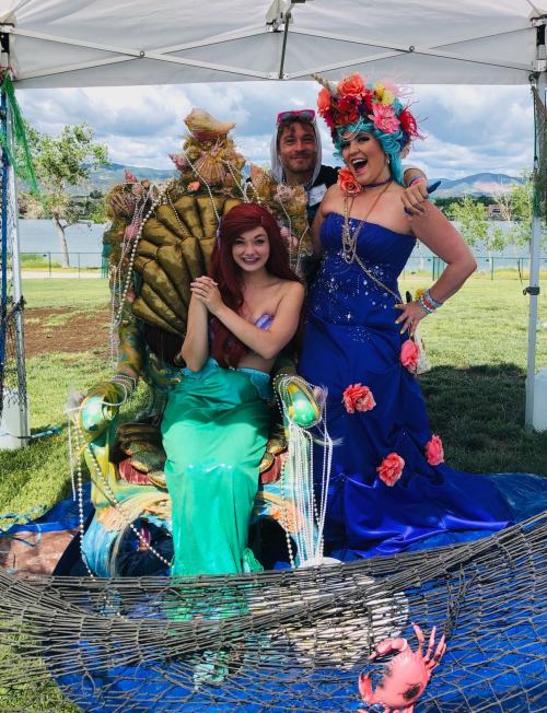 Three people dressed in colorful costumes pose together under a tent by a lake, with a decorative throne and props.