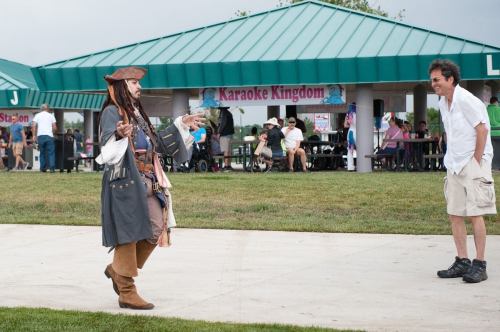 A person dressed as a pirate interacts with a man in a white shirt at an outdoor event with picnic tables.