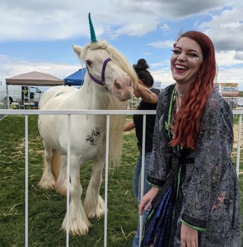 A woman in a colorful outfit laughs beside a white unicorn with a blue horn at an outdoor event.