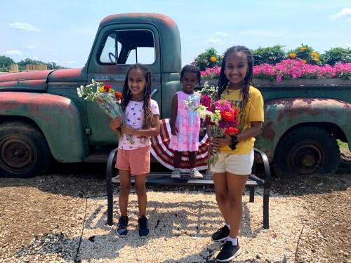 Three girls hold bouquets of flowers in front of a vintage truck adorned with colorful plants.