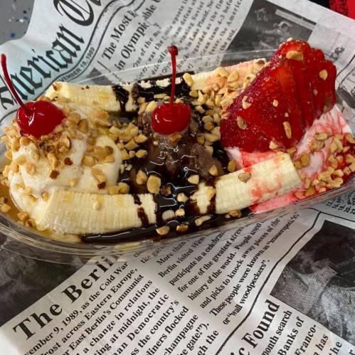 A banana split with scoops of chocolate and strawberry ice cream, topped with whipped cream, nuts, and cherries, on newspaper.