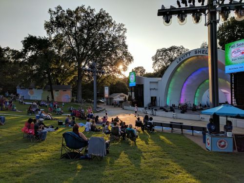 A park scene with people sitting on the grass near a colorful band shell at sunset, enjoying an outdoor concert.