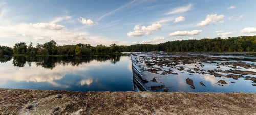 A serene river scene with calm water reflecting clouds and trees, featuring a rocky shoreline and a dam in the distance.