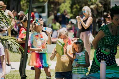 Children at a lively outdoor event, holding crafts and water bottles, with adults and colorful decorations in the background.