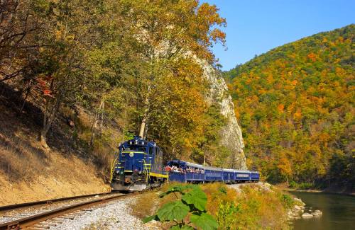 A blue train travels along a scenic route beside a river, surrounded by colorful autumn foliage and hills.