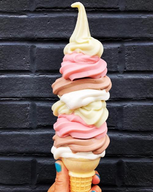 A hand holding a tall ice cream cone with swirls of pink, brown, and white soft serve against a black brick background.