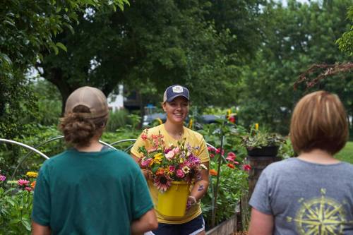 A smiling woman in a yellow shirt holds a bouquet of flowers while talking to two people in a garden.