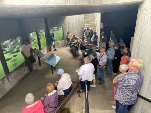 A group of people gathered indoors, listening to a speaker near informational displays about aquatic life.
