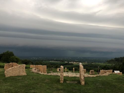 A stone circle on grassy land with dark, stormy clouds looming over a distant landscape.