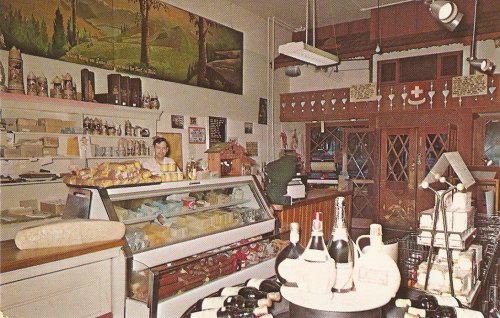 A vintage shop interior featuring a display case of cheeses, wine bottles, and a mural of a scenic landscape.