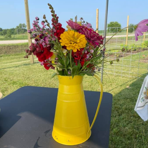 A bright yellow pitcher filled with colorful flowers, including red, pink, and yellow blooms, set on a table outdoors.